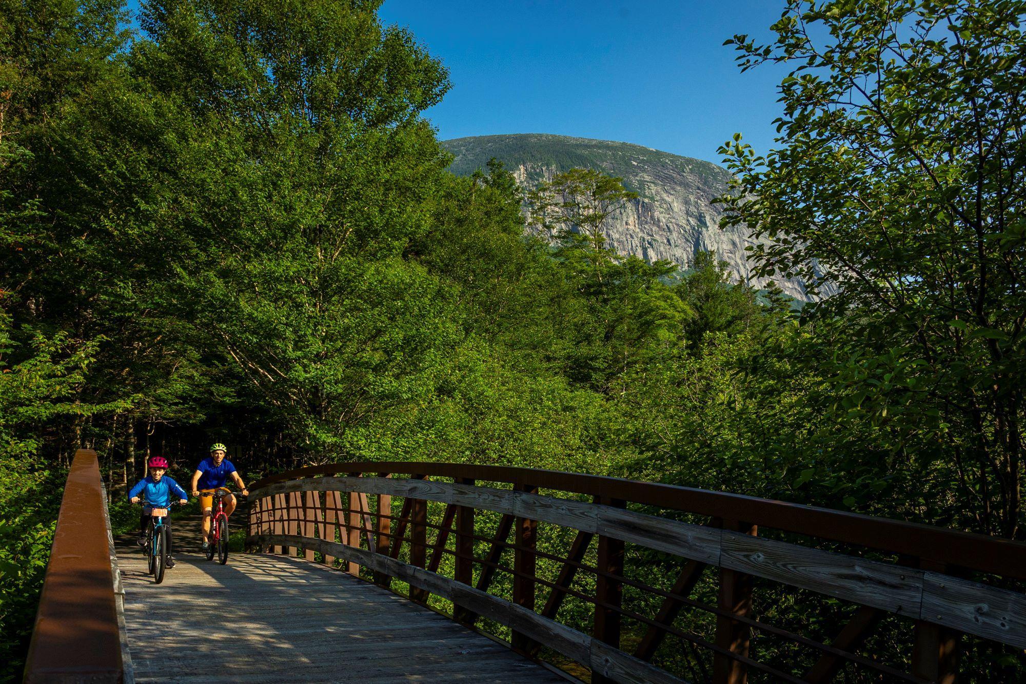 2 bikers cross a bridge with the Cannon Cliff in the background.